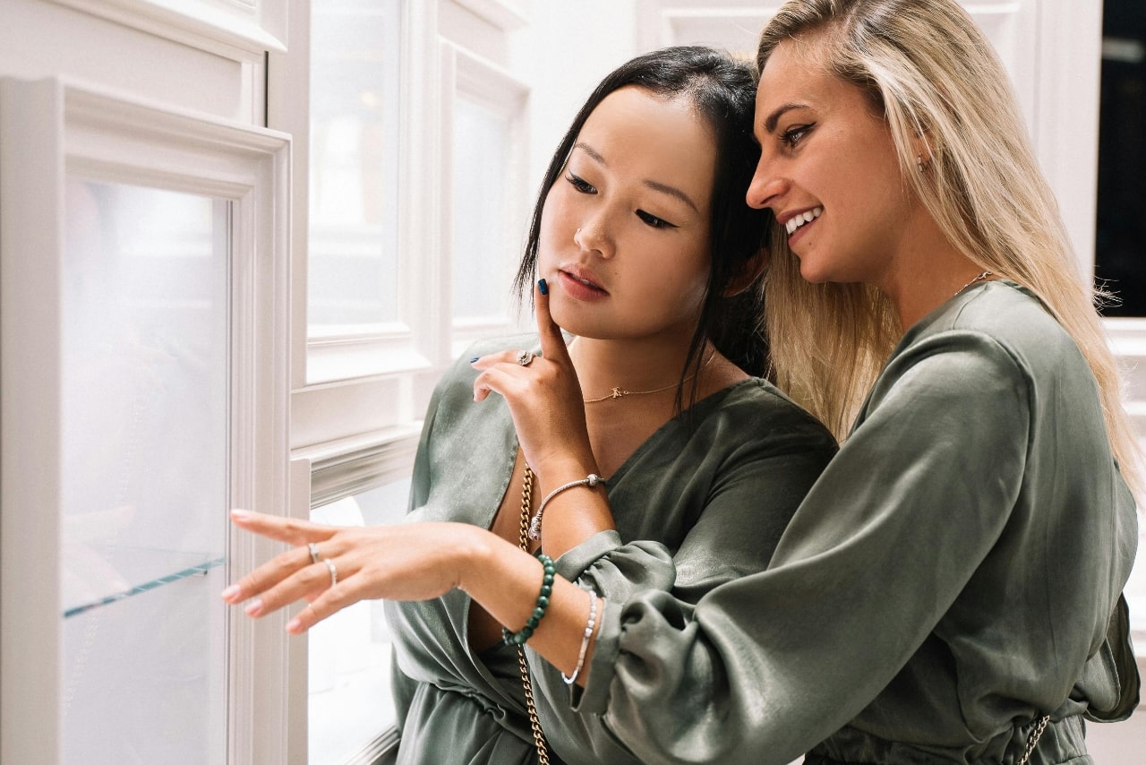 Two women in green dresses admire a jewelry display case, one pointing thoughtfully while the other smiles.