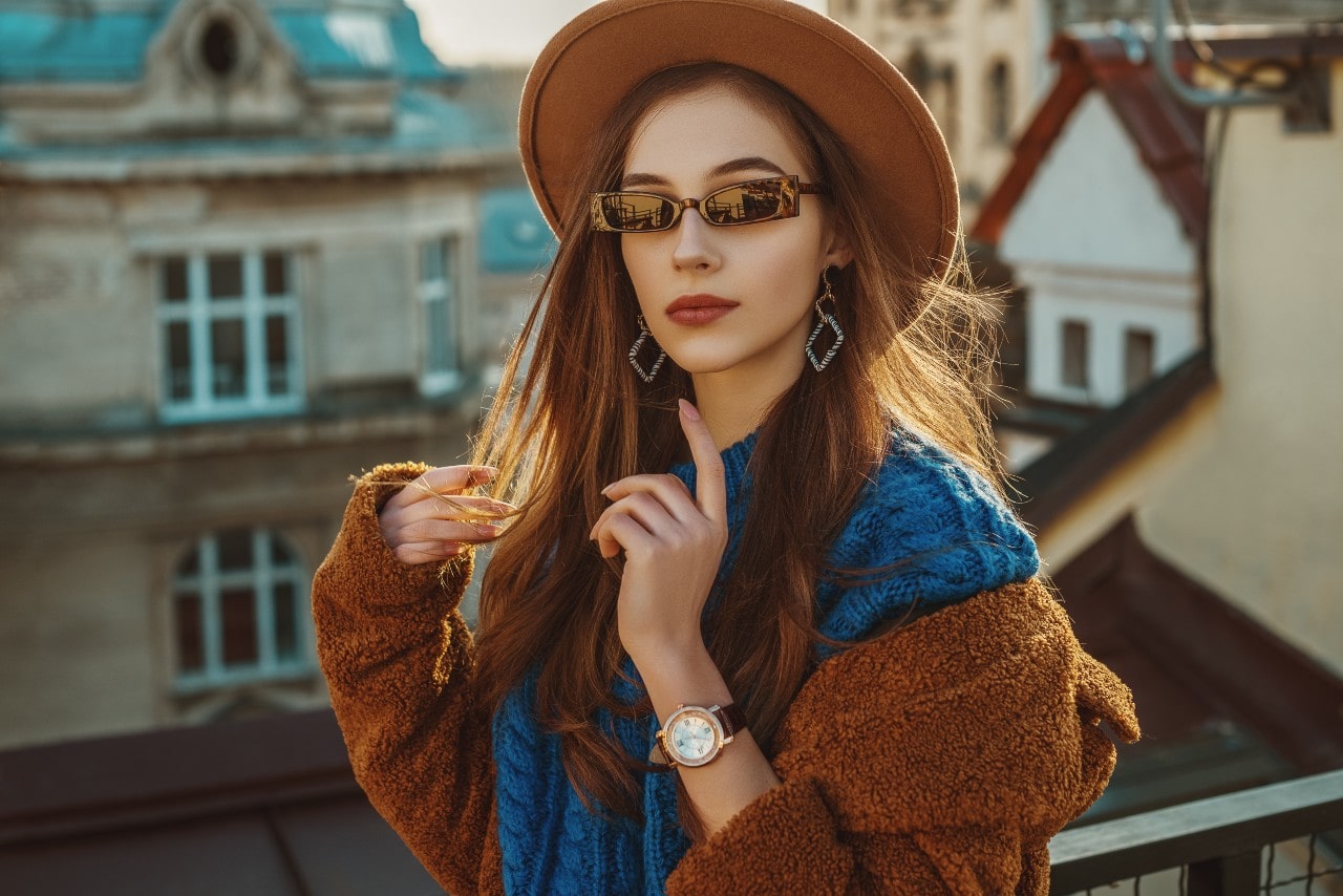 A woman on a rooftop in fall attire, featuring a wide-brimmed hat, sunglasses, blue scarf, and brown coat, set against an urban backdrop.