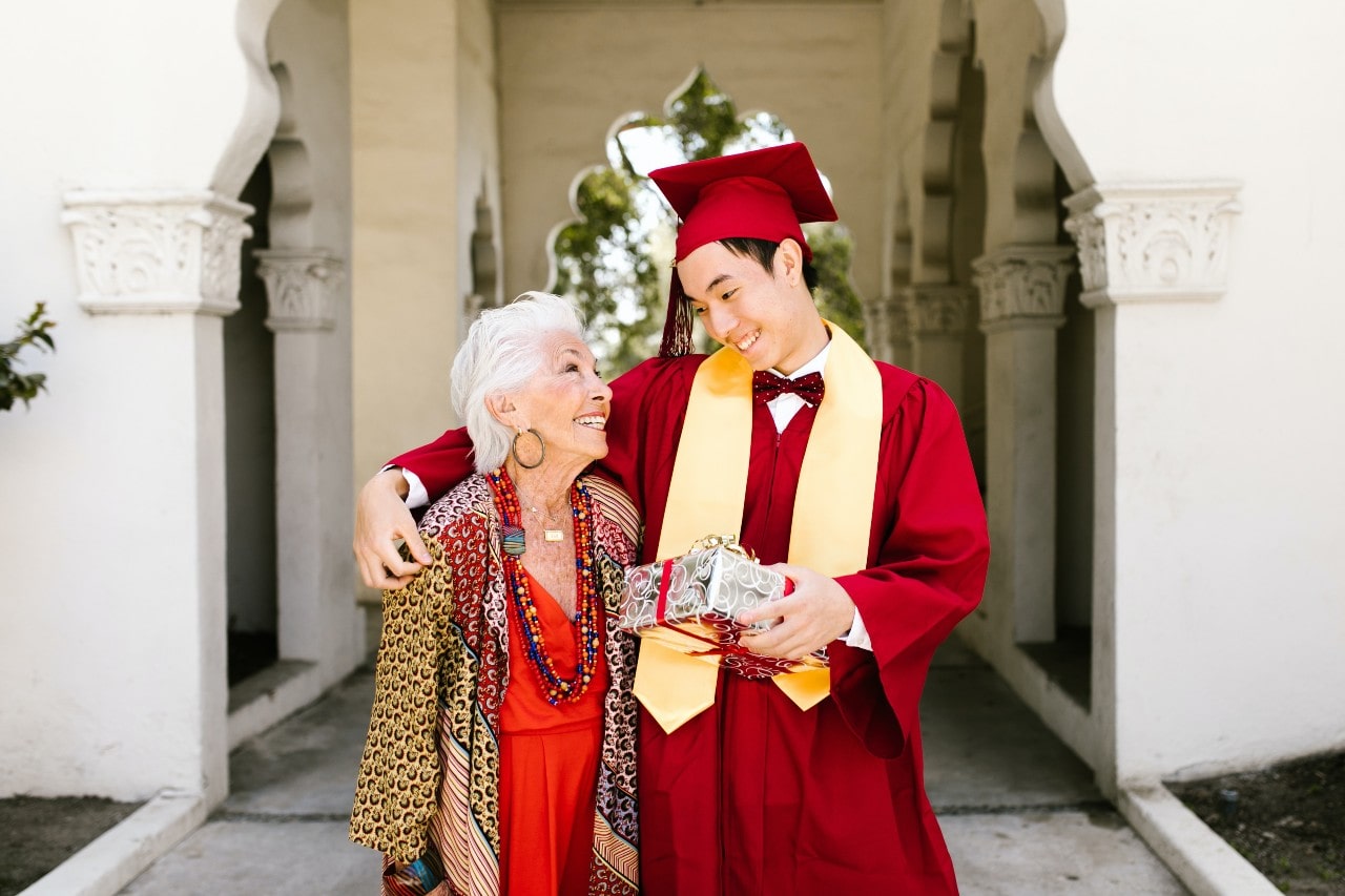 A young man in a red graduation gown and cap holds a wrapped gift and embraces an elderly woman dressed in a red and patterned outfit.