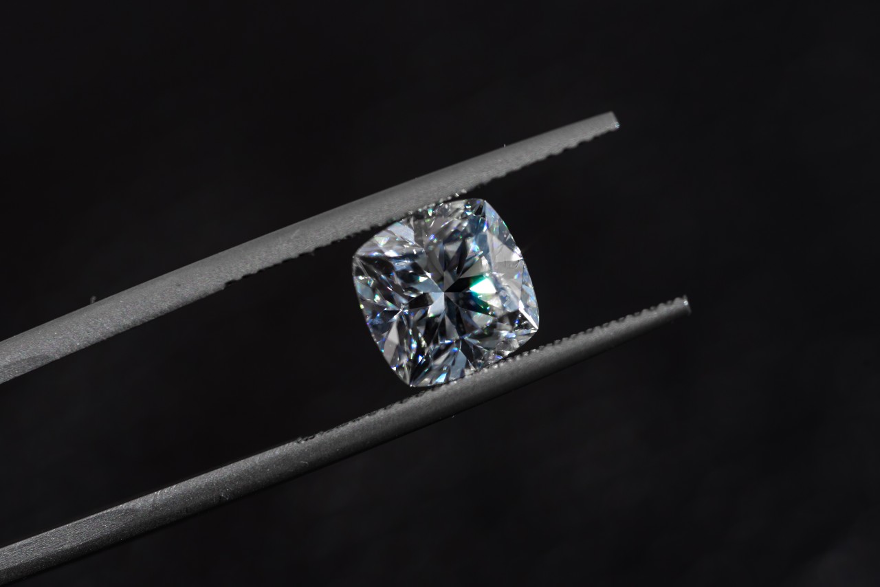 A close-up of a glistening cushion cut diamond held in a pair of jeweler’s tweezers on a black backdrop.