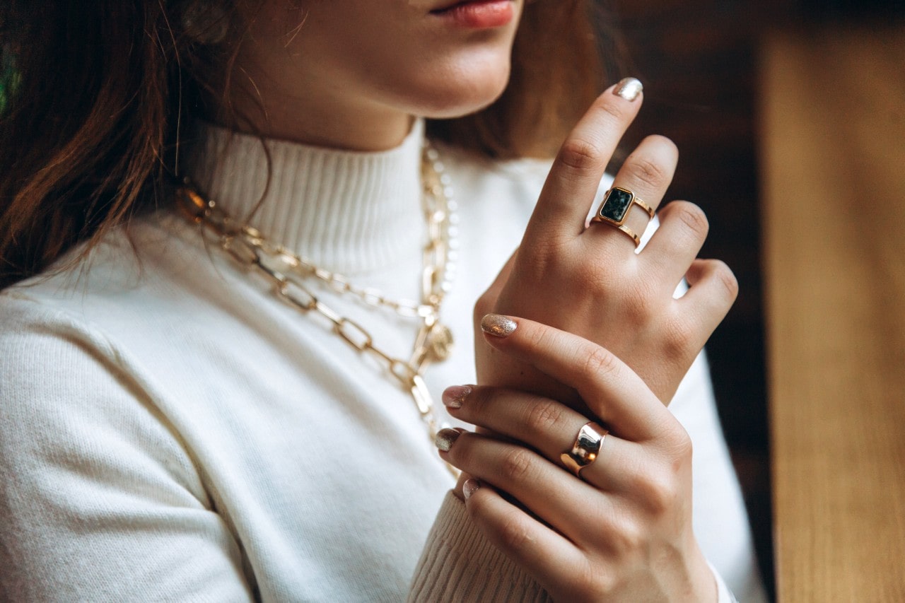 A close-up of a fashionable woman wearing a white turtleneck, her hands adorned with chunky gold rings and her neck adorned with layered chain necklaces.