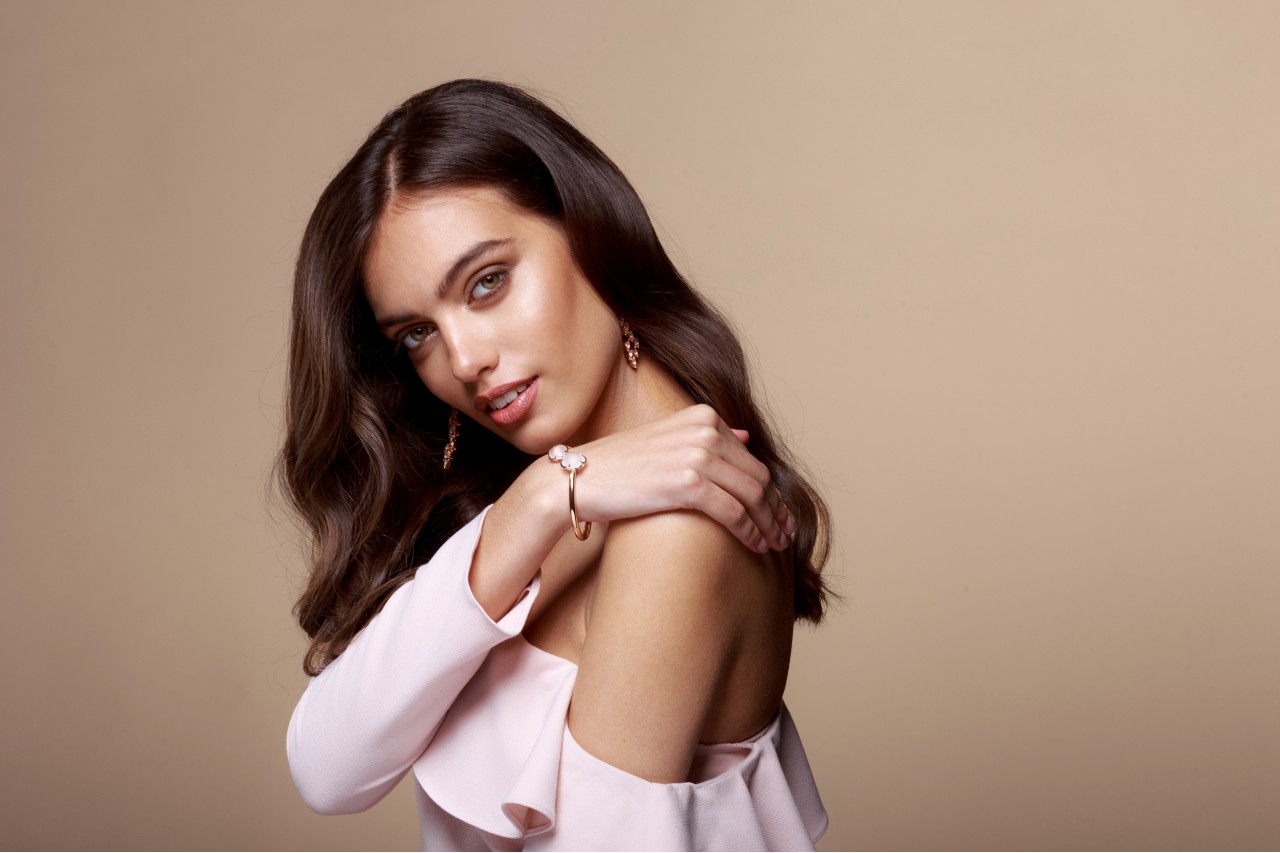 A woman with long brunette hair wearing rose gold earrings and a floral-inspired bracelet, softly resting her hand on her shoulder against a beige background