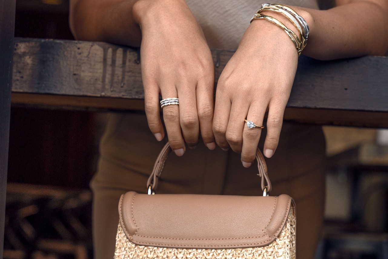 A close up of a woman's hand, adorned with stacked rings and gold bangles, holding a brown leather bag, leaning on a wooden surface.