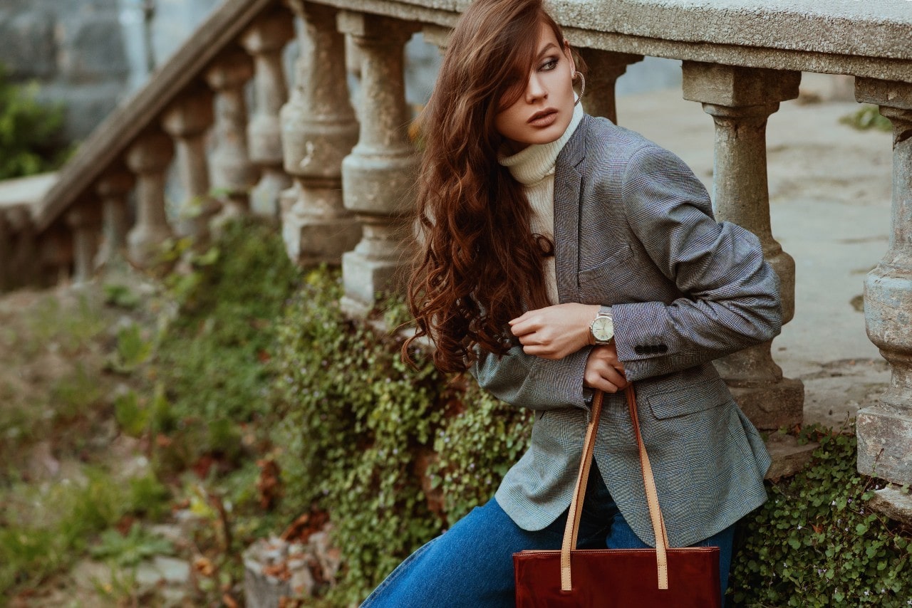 A woman in a gray blazer and turtleneck sits by a stone balustrade, holding a brown tote.