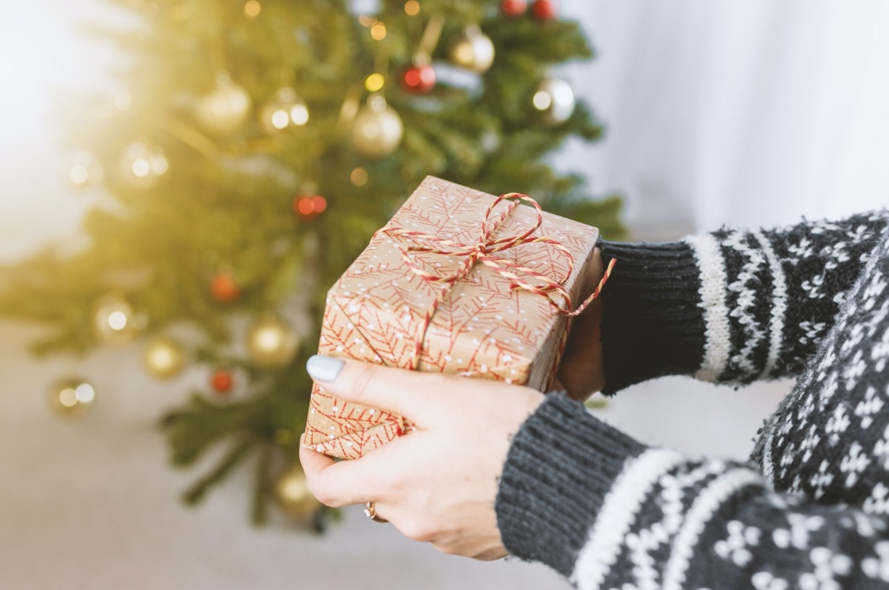 A close up of a hands in a grey sweater holding a wrapped gift in front of blurred christmas tree
