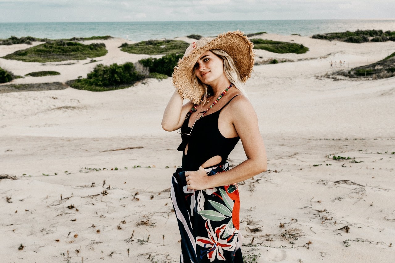 A woman in vacation clothing and a big straw hat poses for the camera, lots of sand and the ocean shore behind her.
