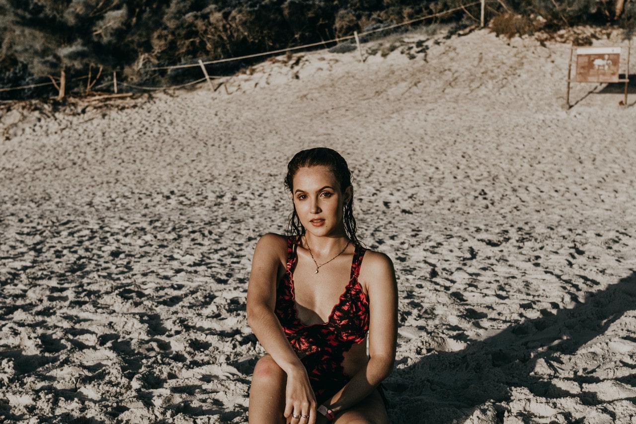 A woman in a red and black bathing suit sitting on sand and posing for the camera.
