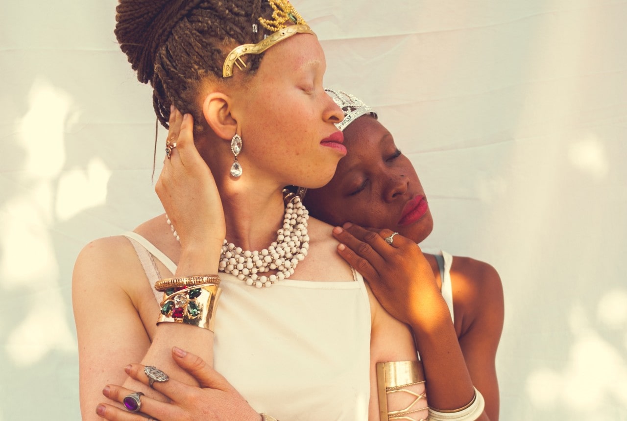 Two women embracing, adorned in jewelry and white clothing, against a white background.