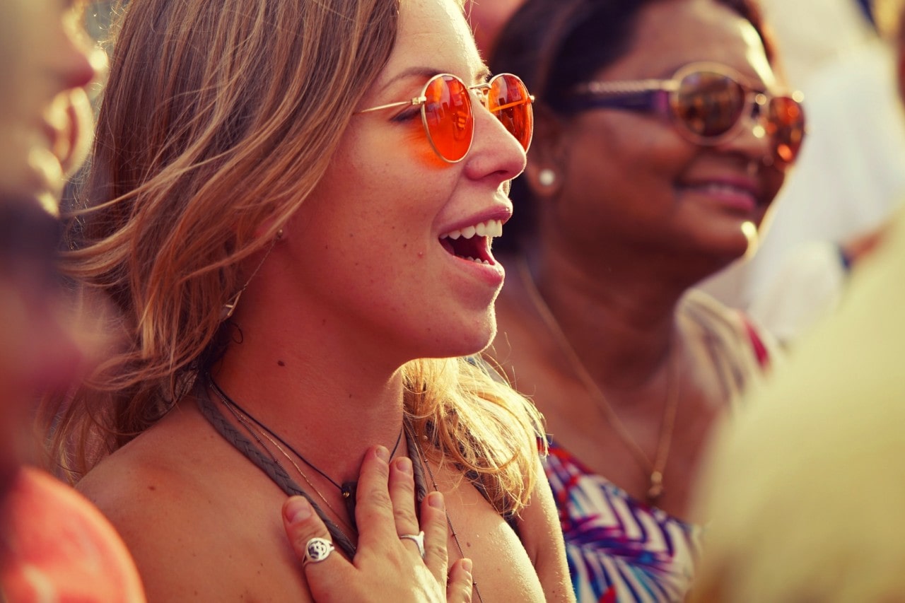 A woman with orange-tinted sunglasses singing with emotion at an outdoor event, surrounded by other smiling attendees.