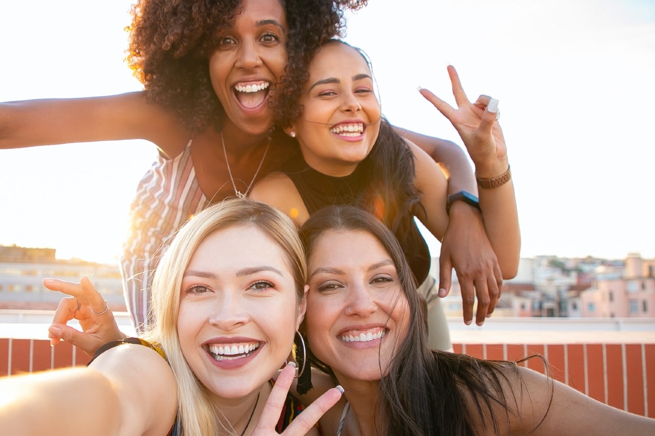 A group of four young women smiling and posing joyfully for a selfie on a rooftop during golden hour.