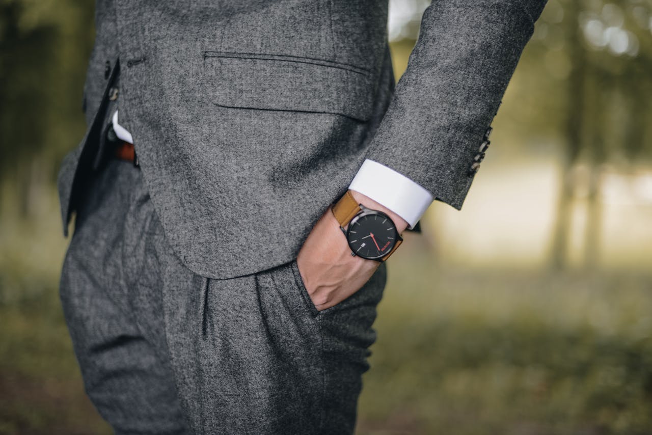 A view of a well-dressed man in a suit standing in a lightly wooded area, with emphasis on the luxury watch on his left wrist.