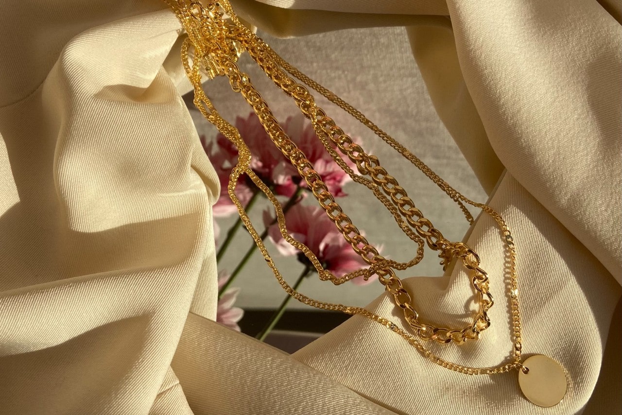 A close-up of yellow gold chain necklaces on a mirror and reflecting pink flowers in the background.
