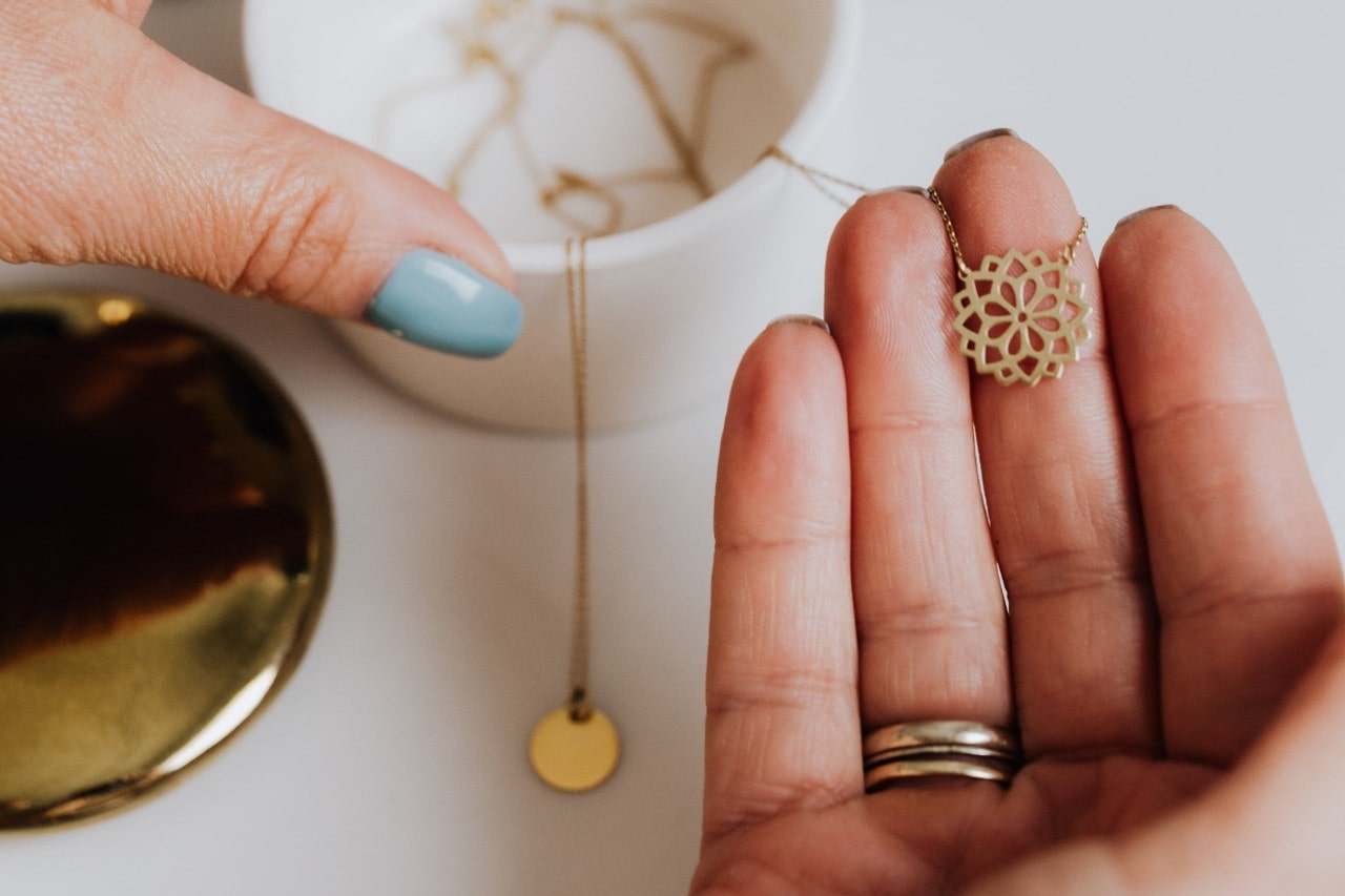 A close-up of a person holding a pendant of a yellow gold necklace with another one laying on a table in the background.
