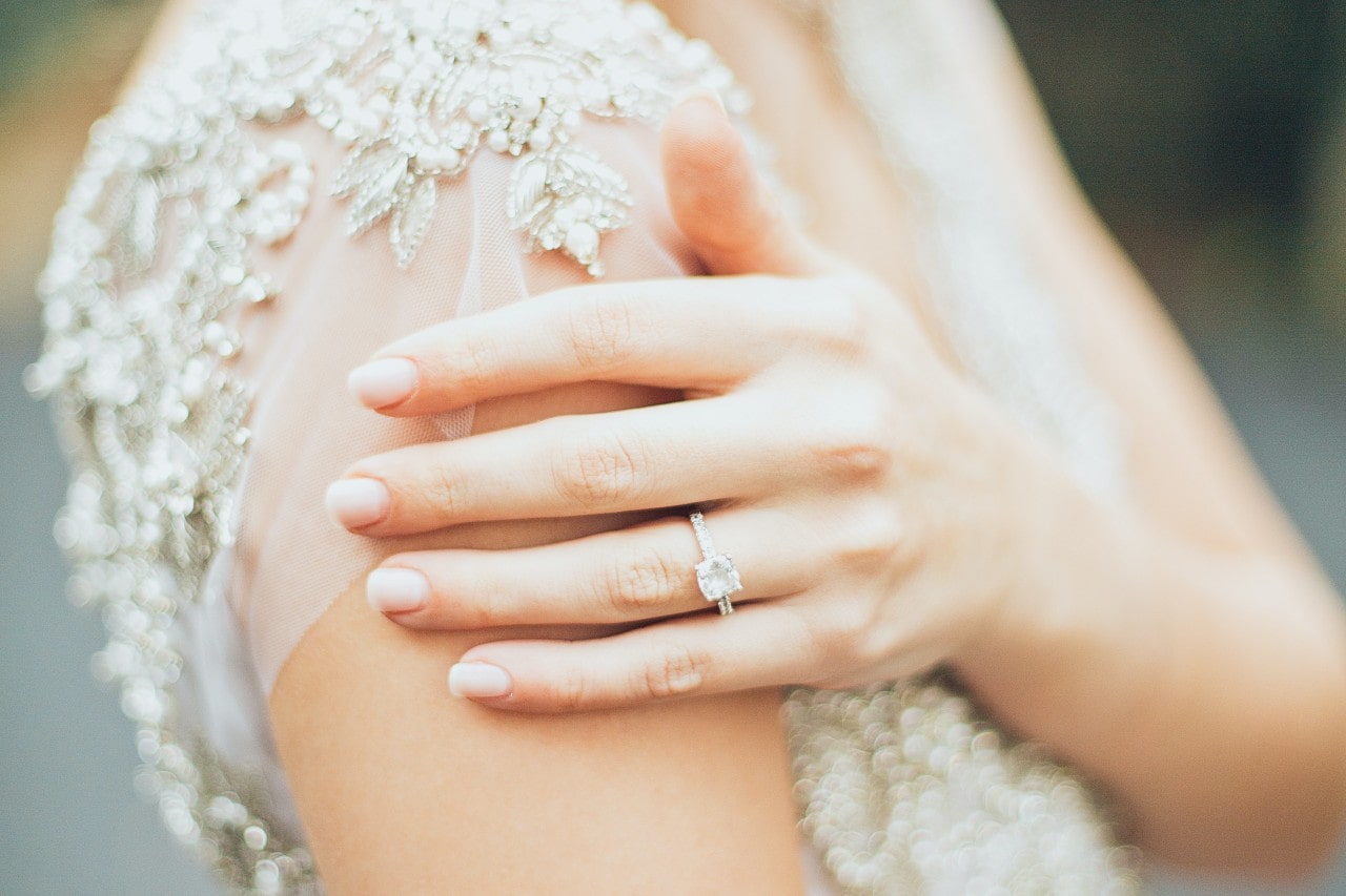 Close-up of a woman’s hand with a side stone engagement ring resting on a shoulder adorned with intricate jeweled embroidery.