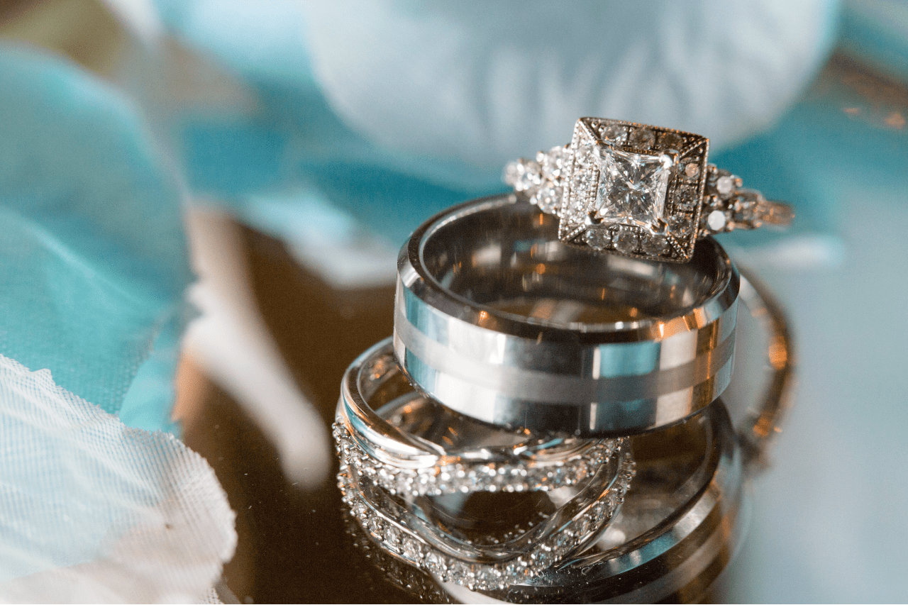 A close-up of three wedding rings on a reflective surface: diamond-studded engagement ring atop two bands, with blurred blue fabric in the background.