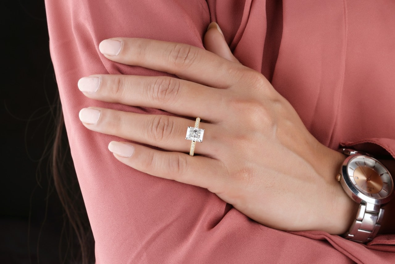 A woman wearing a pink shirt and a gold emerald cut engagement ring with side stones
