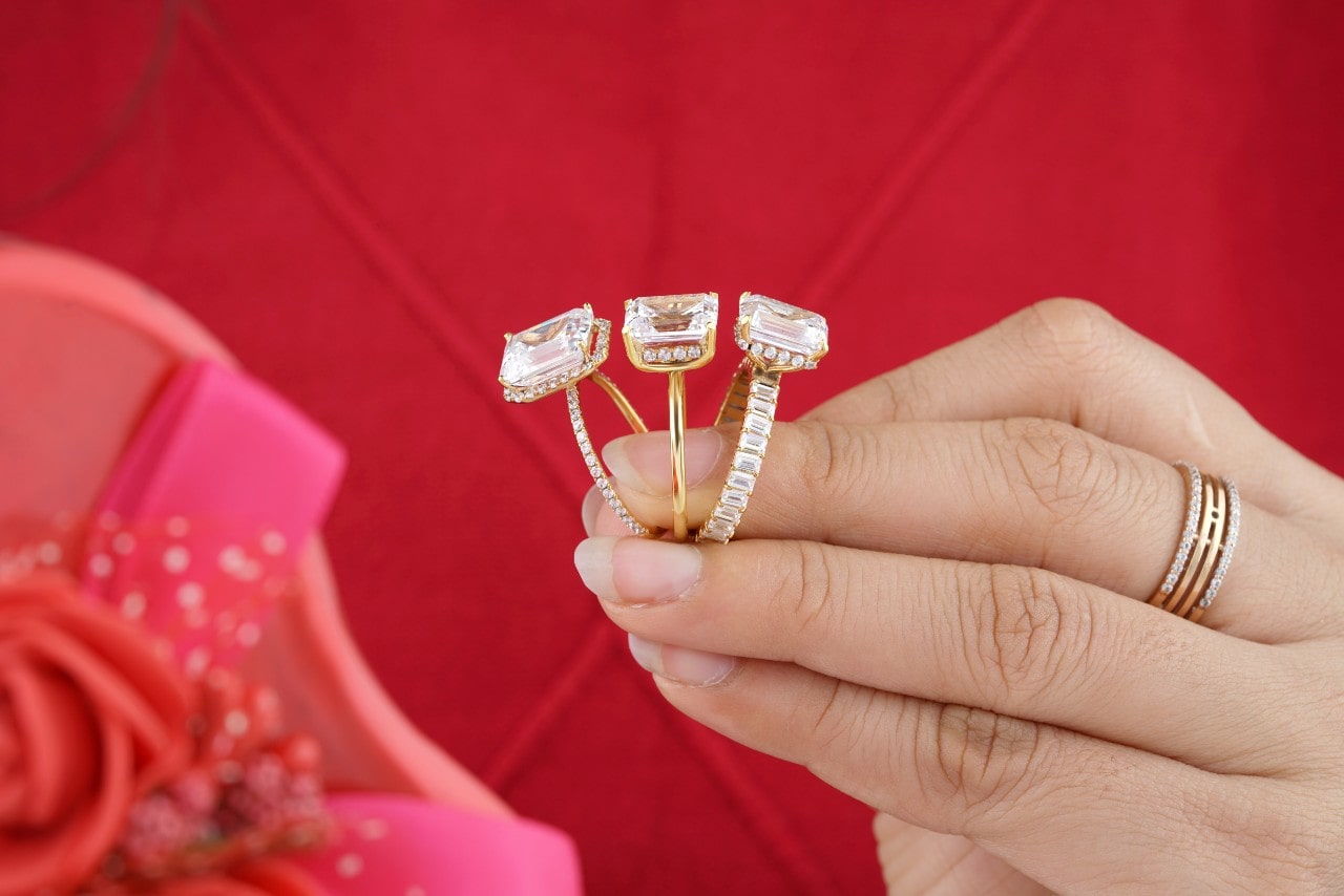 Hand holding three emerald cut engagement rings against a red background