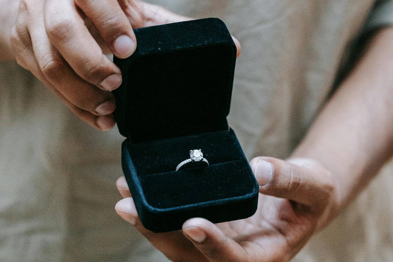 A person holding an open black velvet box displaying a side stone diamond engagement ring.