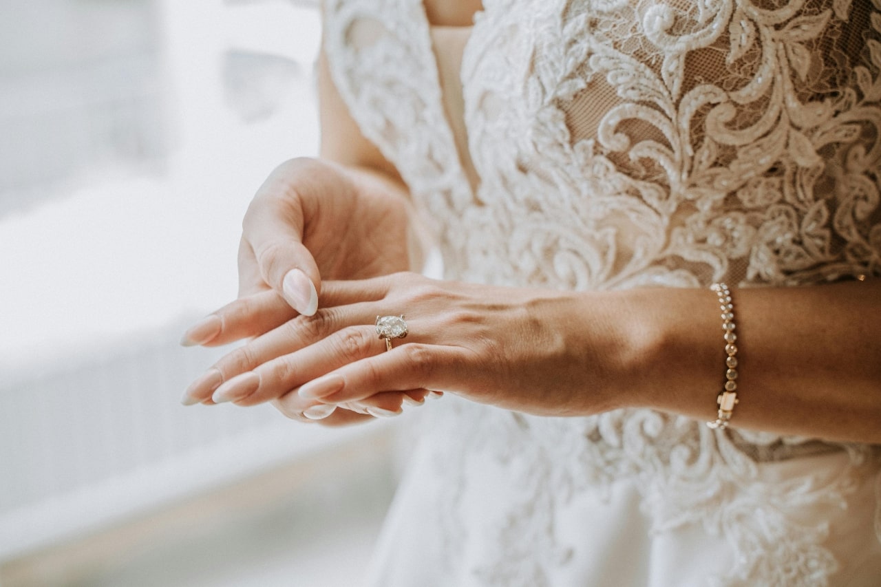 A close-up of a bride in a lace wedding gown, an engagement ring with massive oval cut diamond on her finger.