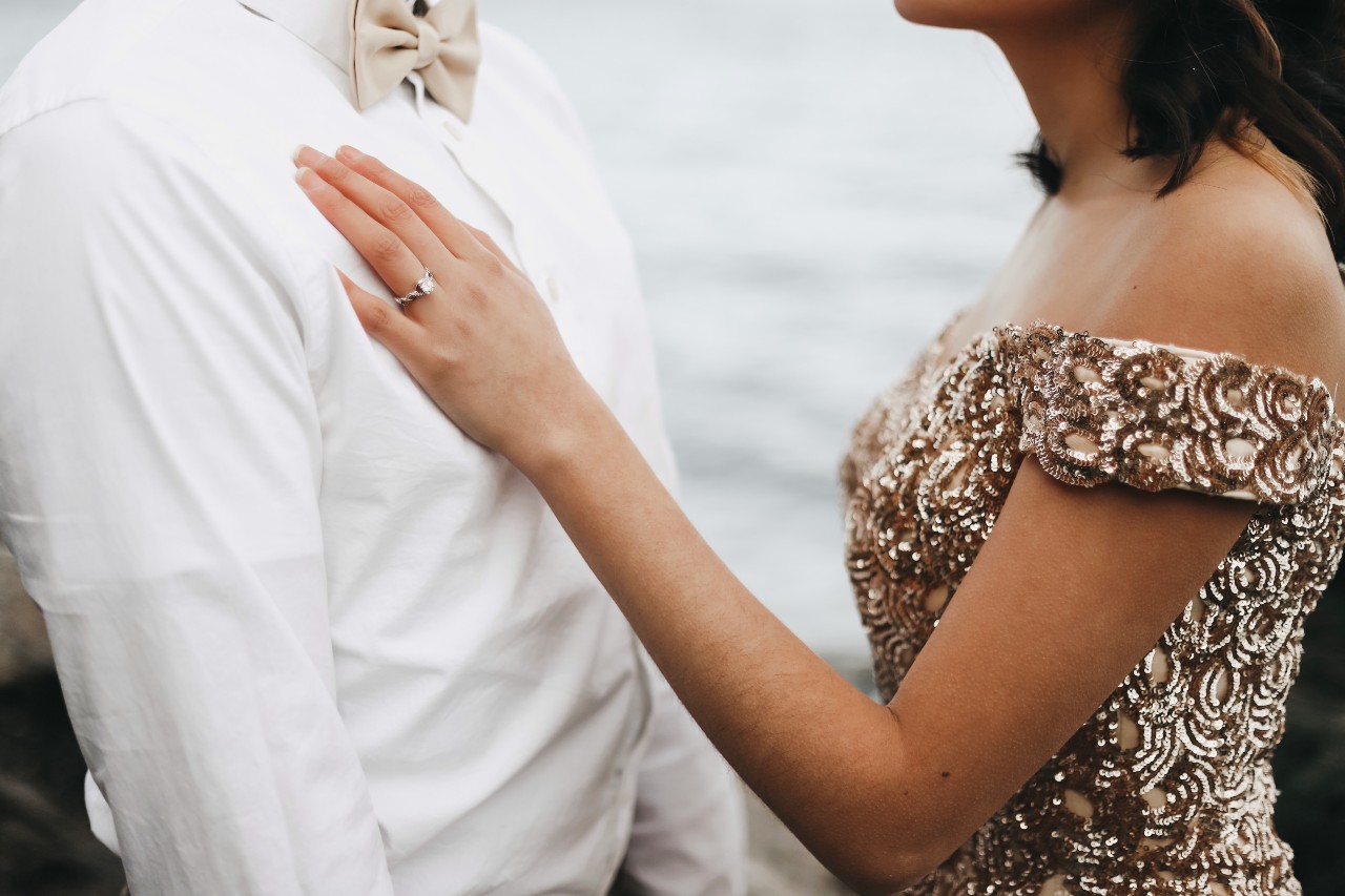 A woman in a sparkling yellow gold dress put her hand on a man in white shirt, showcasing a rose gold diamond engagement ring. A woman in a sparkling yellow gold dress put her hand on a man in white shirt, showcasing a rose gold diamond engagement ring.