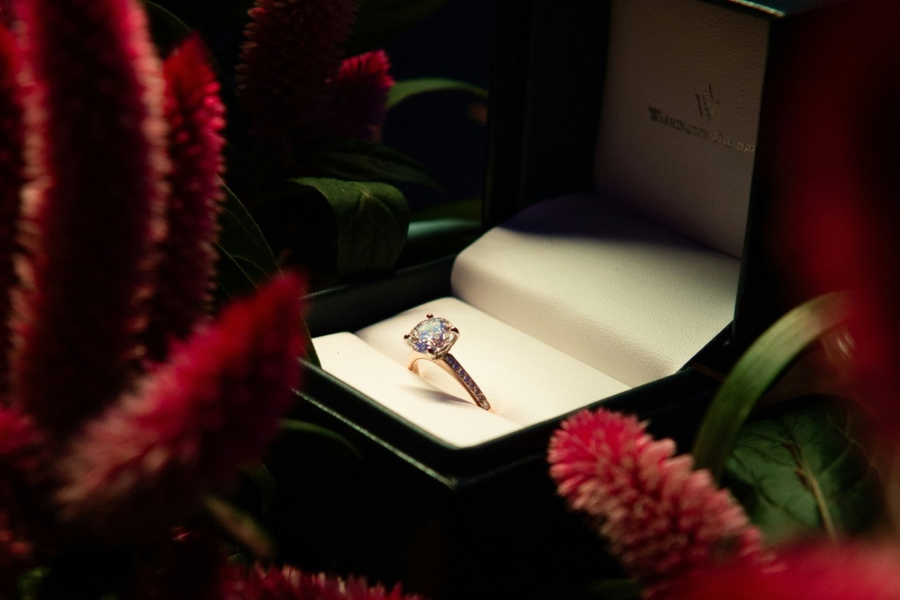 A close-up of a round cut, side stone engagement ring in a green ring box surrounded by flowers. A close-up of a round cut, side stone engagement ring in a green ring box surrounded by flowers.