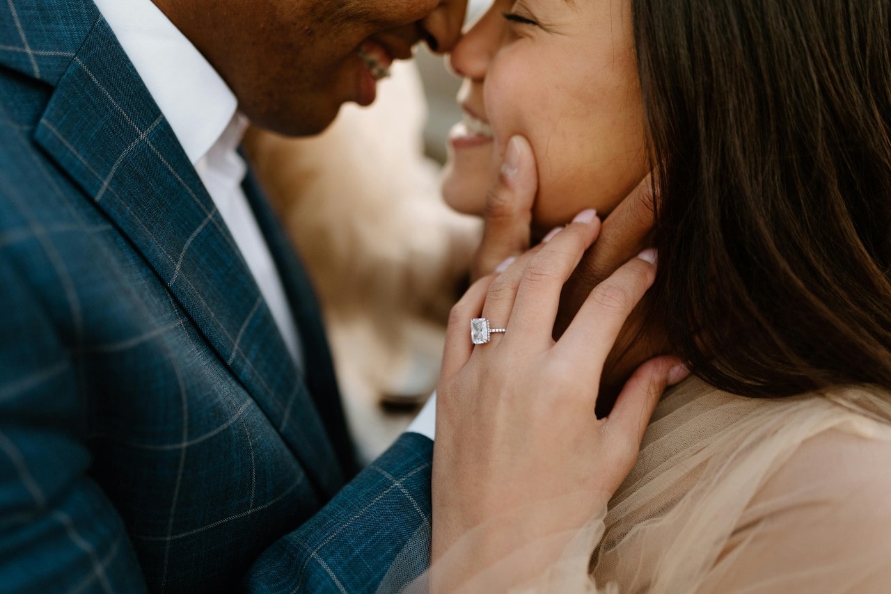 A close-up of a man and woman smiling at each other— the woman wearing an emerald cut engagement ring A close-up of a man and woman smiling at each other— the woman wearing an emerald cut engagement ring