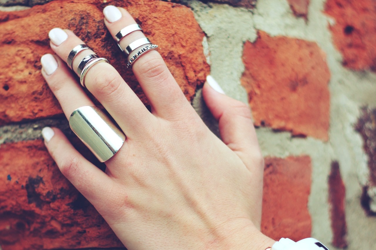 A hand with white nail polish rests on a brick wall, adorned with multiple silver rings of varying designs, creating a stylish and edgy look.