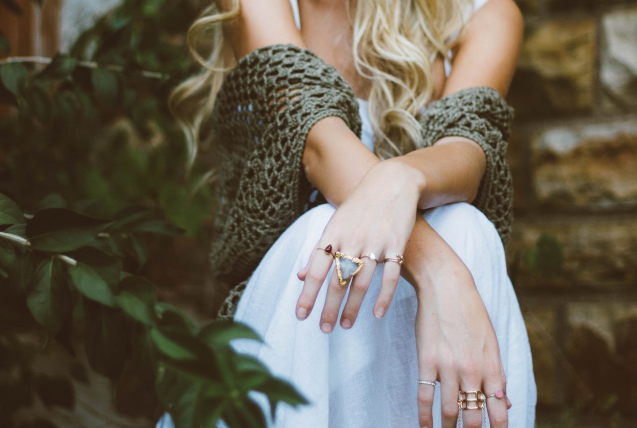 A lady with long blonde hair sits with their arms crossed, wearing a green crochet shawl, white skirt, and multiple rings.