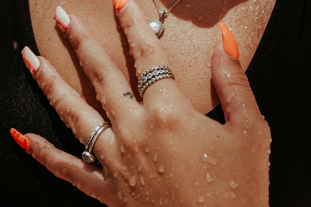 Close-up of a hand with colorful nails, adorned with various rings, resting on a wet chest