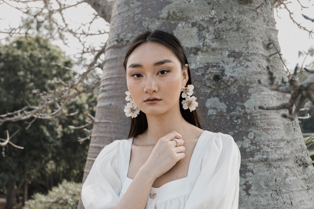 A view of a young woman wearing a breezy white top and large floral earrings, standing in front of a tree. A view of a young woman wearing a breezy white top and large floral earrings, standing in front of a tree.