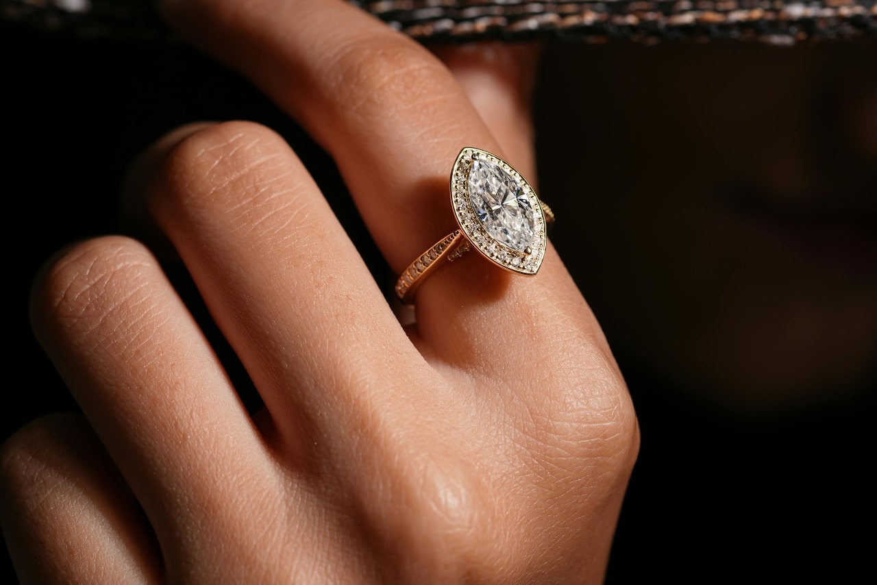 A close up of a woman’s finger, showcasing yellow gold marquise cut diamond engagement ring against a dark blurry background. A close up of a woman’s finger, showcasing yellow gold marquise cut diamond engagement ring against a dark blurry background.