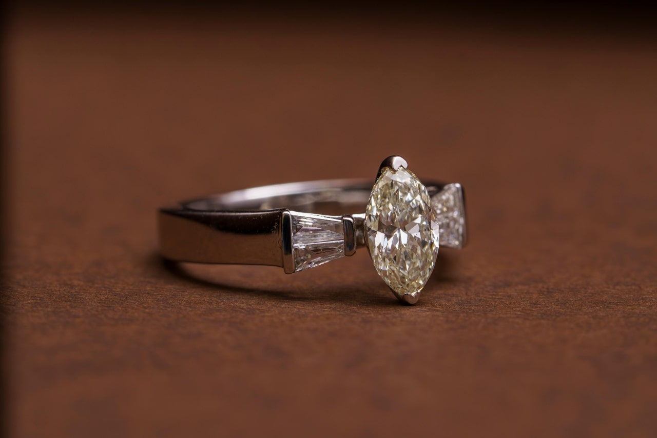 A close up of a silver three stone engagement ring with a marquise cut diamond as its center stone placed on a blurry brown surface. A close up of a silver three stone engagement ring with a marquise cut diamond as its center stone placed on a blurry brown surface.