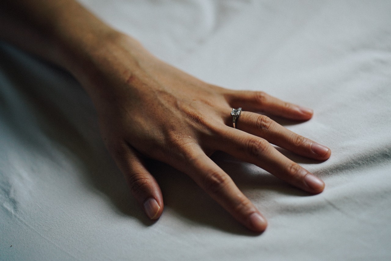 A woman’s hand resting on a white sheet, wearing a princess cut engagement ring