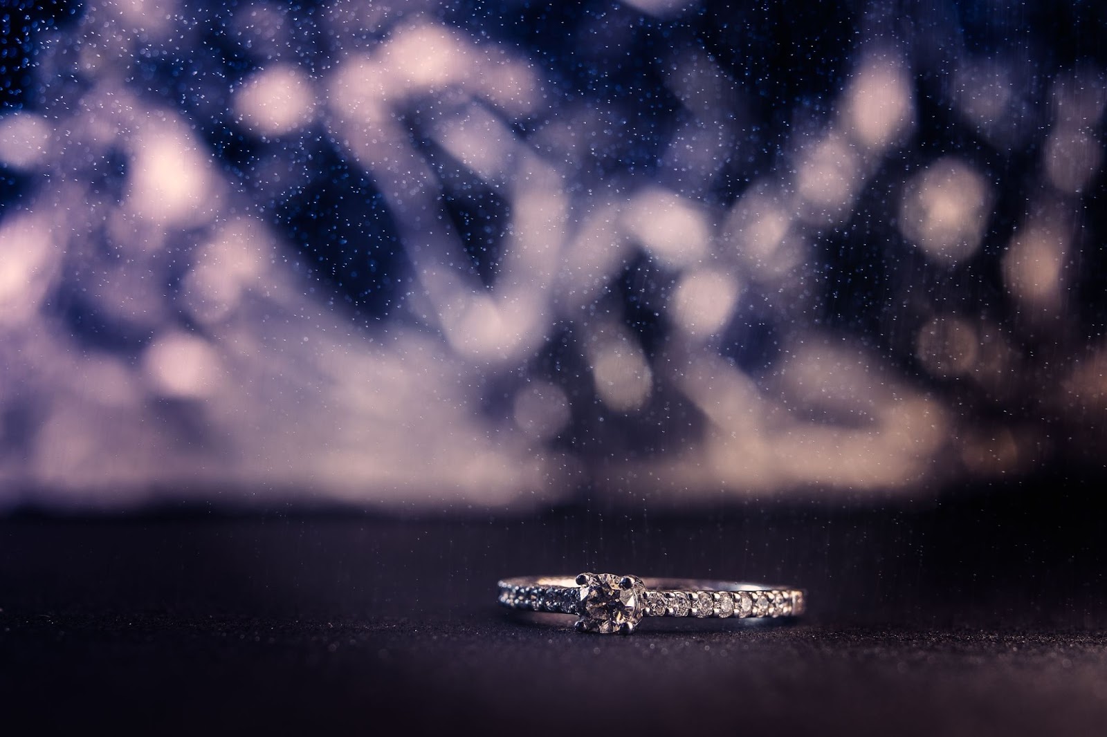 Side stone engagement ring lying on a dark surface with glittering white lights in the background
