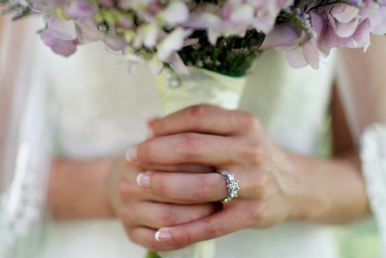 Bride holding a bouquet of purple flowers, with a close-up on her three stone diamond engagement ring