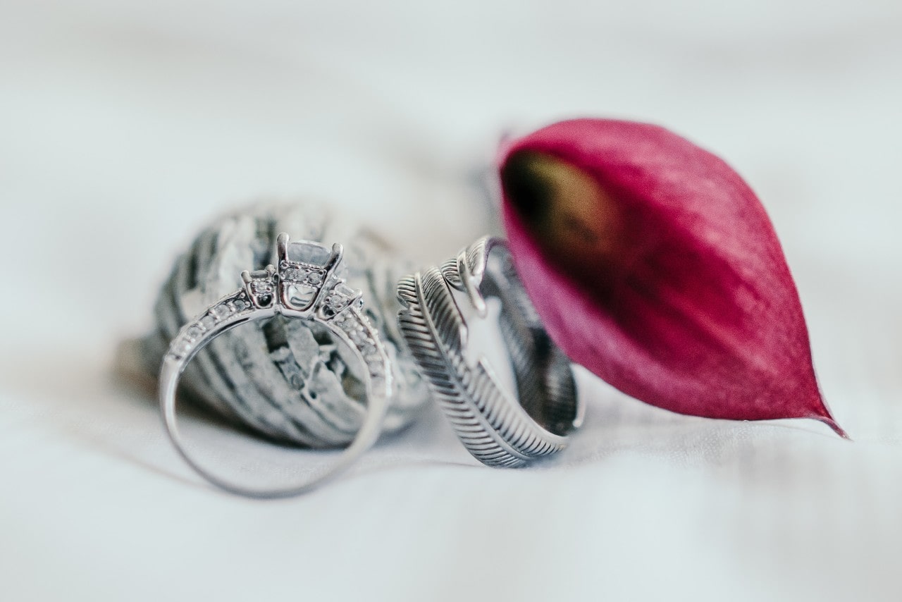 A side profile of a three stone ring with a feather motif band in front of a red flower and seashell