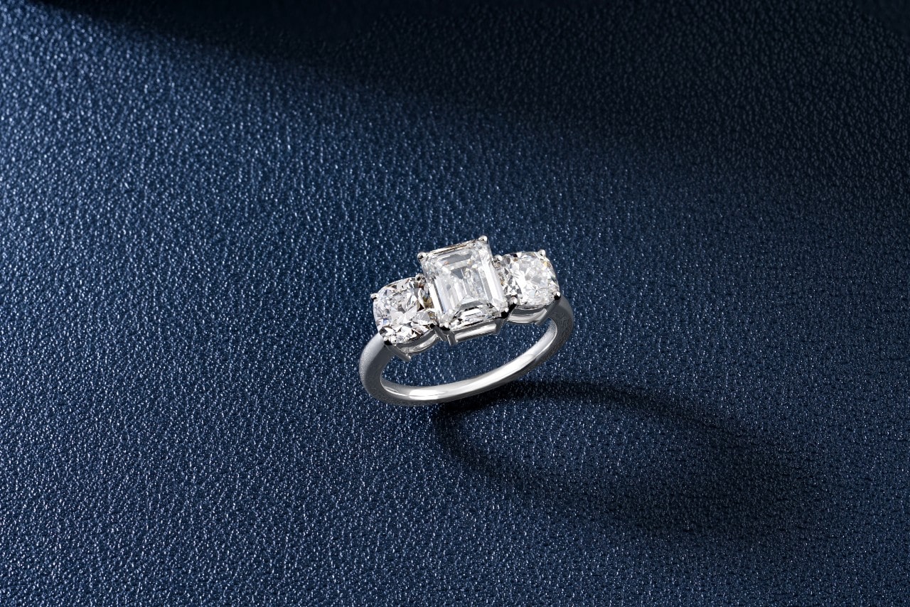 A three stone cushion cut engagement ring on a navy blue leather background