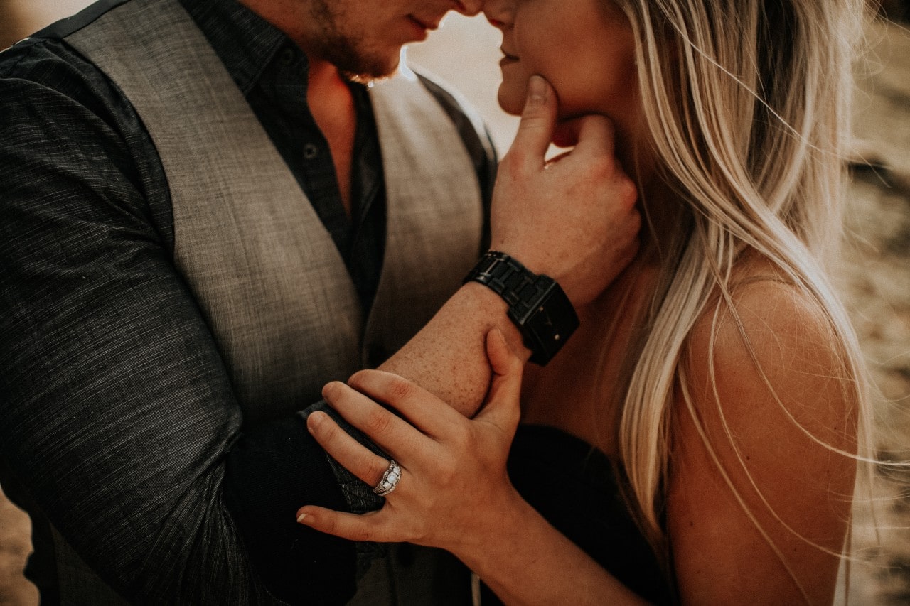 A couple nose to nose with the woman’s hand showing the thick band of an engagement ring is resting on the man’s rest as he rests a thumb on her face