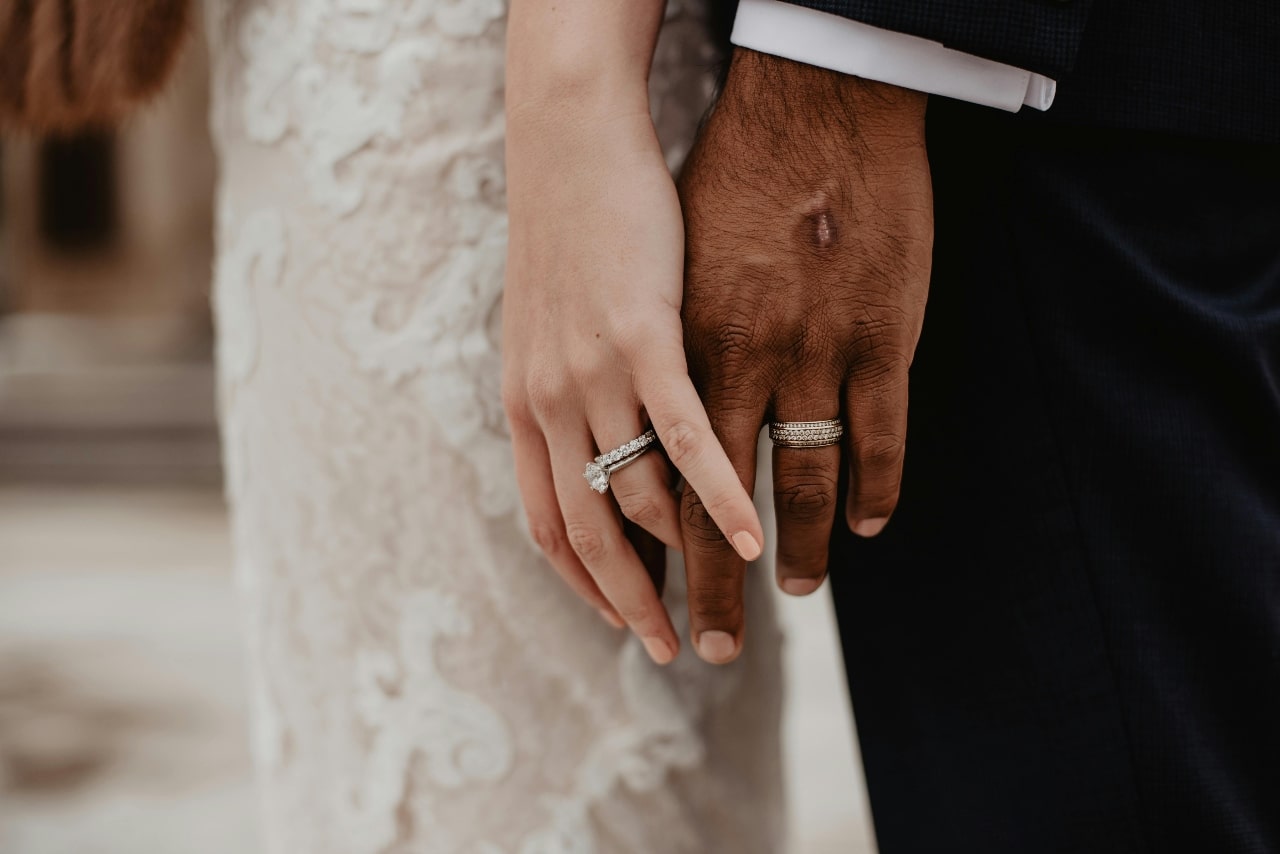 A bride and groom standing side by side, showing their matching wedding rings on clasped hands.