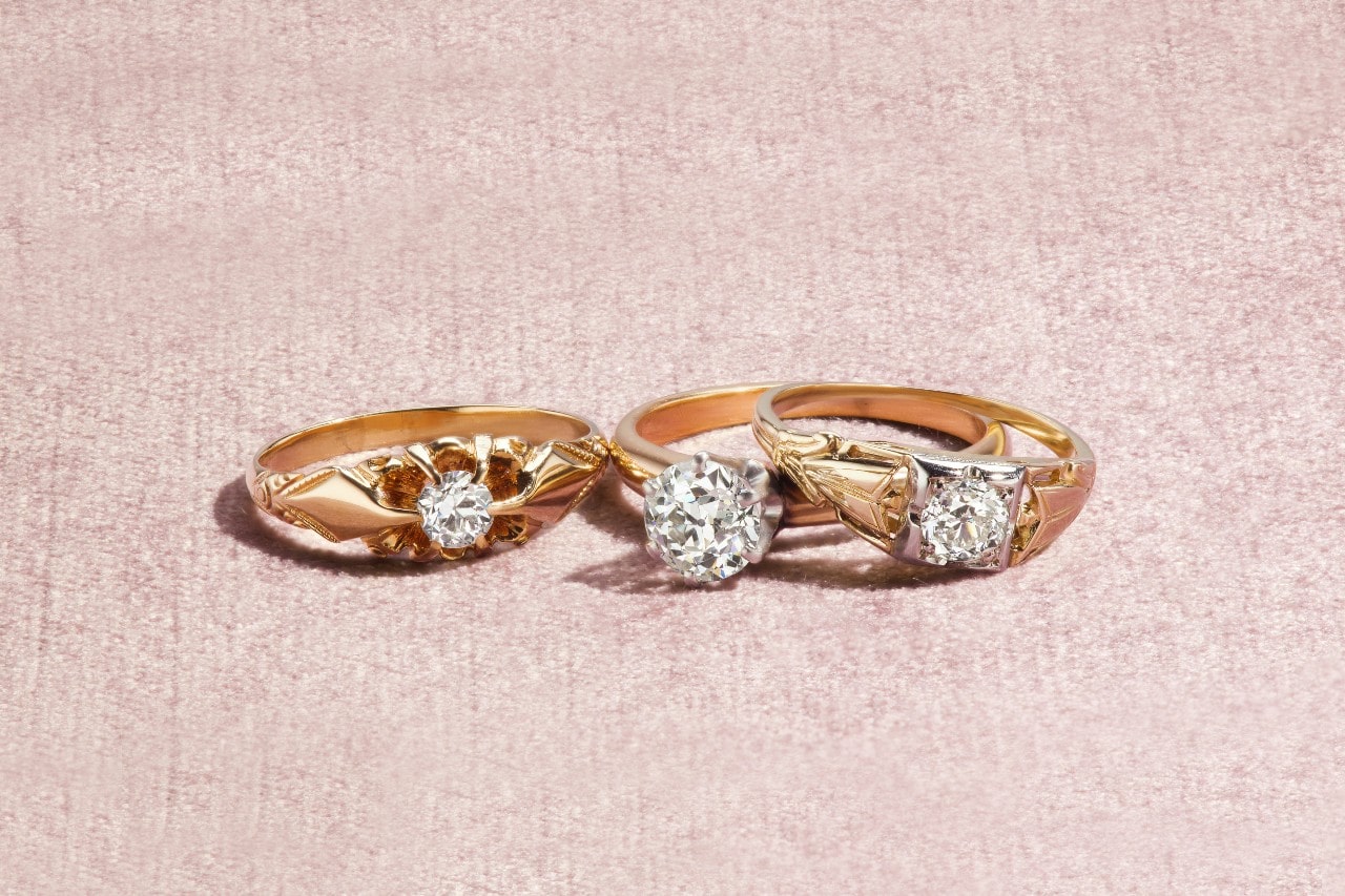 Three vintage-style gold rings with prominent diamonds displayed on a soft pink surface