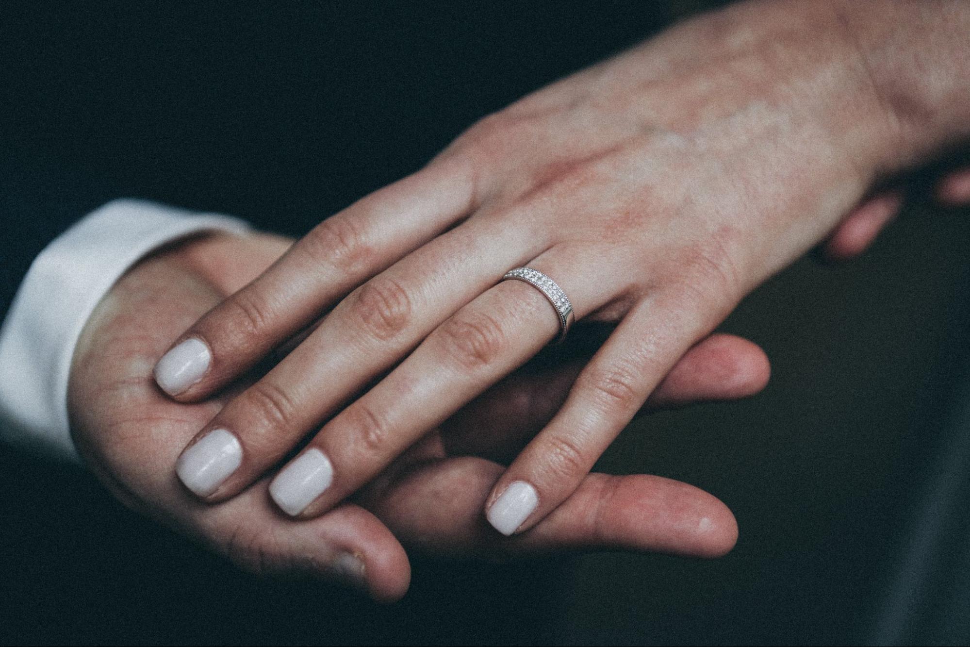 A close-up of a person’s hand wearing a diamond-encrusted wedding band gently held by another hand