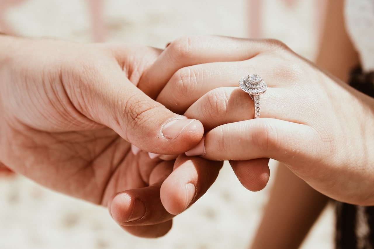 A halo engagement ring sits on a woman’s finger as her partner holds her hand.