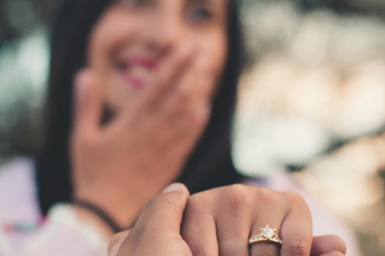A close-up of a woman’s hand with a diamond engagement ring, her happy and tearful face in the background.